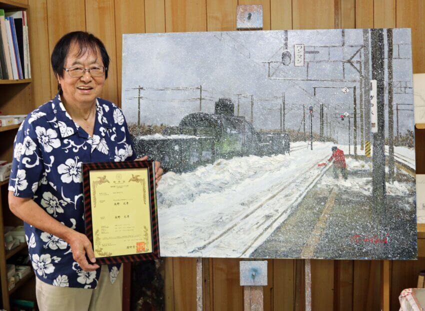 作品の前で特許証を手にする高野画伯